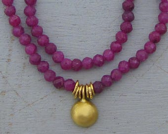 May include: A double-strand necklace featuring small, faceted, deep magenta-colored beads. The necklace includes a central gold-toned pendant shaped like a smooth, rounded disc, suspended from a series of small, vertical gold-toned metal rings.