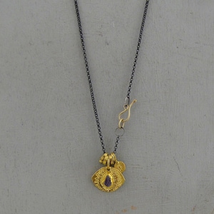 May include: A necklace featuring a dark oxidized silver chain with a gold-toned hook clasp. The pendant is a textured gold-toned circular disc with a small purple stone at the center and a tiny gold-toned sun charm attached to the top bail.