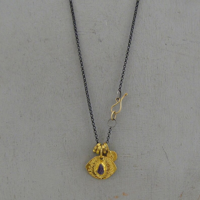 May include: A necklace featuring a dark oxidized silver chain with a gold-toned hook clasp. The pendant is a textured gold-toned circular disc with a small purple stone at the center and a tiny gold-toned sun charm attached to the top bail.