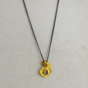 May include: A necklace featuring a dark, oxidized silver-toned chain and a gold-toned pendant. The pendant consists of two layered, textured circular discs with a small, deep purple, teardrop-shaped stone set in the center of the front disc.