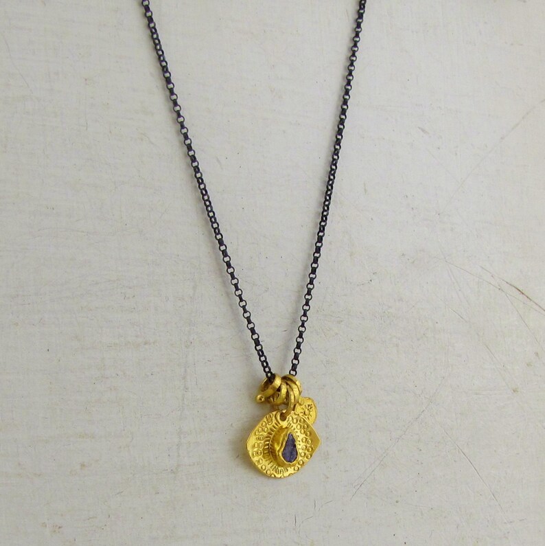 May include: A necklace featuring a dark, oxidized silver-toned chain and a gold-toned pendant. The pendant consists of two layered, textured circular discs with a small, deep purple, teardrop-shaped stone set in the center of the front disc.