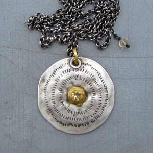 May include: A silver necklace with a round pendant featuring a circular pattern and a small, clear gemstone set in a gold center.