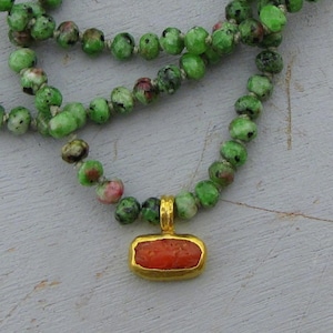 May include: A necklace featuring a strand of round, mottled green and black beads with a central rectangular pendant. The pendant has a textured, bright orange stone inlay set within a polished gold-toned metal frame with a small top loop.