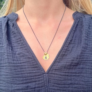 May include: A necklace featuring a dark, oxidized silver chain and a gold-toned pendant. The pendant consists of a textured, irregular gold-colored disc with a small, teardrop-shaped blue stone set in the center and a tiny gold circular charm.