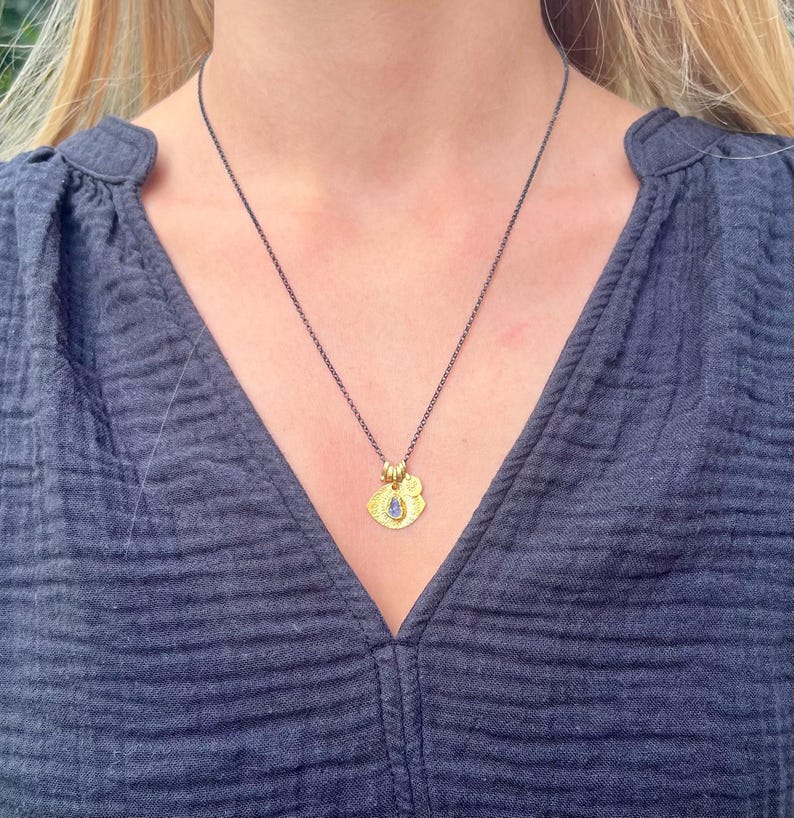 May include: A necklace featuring a dark, oxidized silver chain and a gold-toned pendant. The pendant consists of a textured, irregular gold-colored disc with a small, teardrop-shaped blue stone set in the center and a tiny gold circular charm.