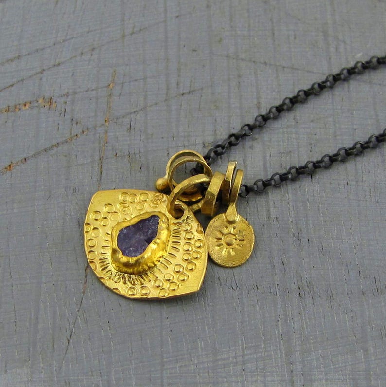 May include: A necklace featuring a dark oxidized silver chain and a gold-toned pendant. The pendant consists of a textured, fan-shaped piece with a central raw purple stone and a small, round charm embossed with a sun design attached to the bail.