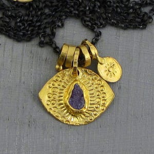 May include: A gold-toned pendant necklace featuring a central, irregular purple stone set within a textured, eye-shaped medallion. The medallion has a sunburst pattern and is suspended from a dark, oxidized chain with a small, round charm.