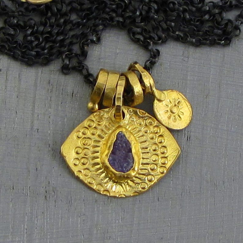 May include: A gold-toned pendant necklace featuring a central, irregular purple stone set within a textured, eye-shaped medallion. The medallion has a sunburst pattern and is suspended from a dark, oxidized chain with a small, round charm.