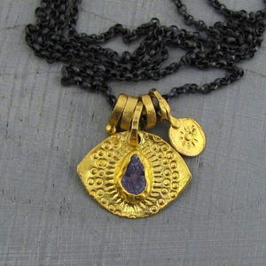 May include: A necklace featuring a dark oxidized silver chain and a gold-toned pendant. The pendant has a textured, circular design with a central, irregular purple stone and a small, separate gold-toned charm embossed with a sun symbol.