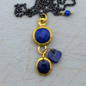 May include: A gold-toned necklace with a dark chain features three blue lapis lazuli pendants. The pendants are round and square, with gold-colored bezels. A small blue bead accents the chain.