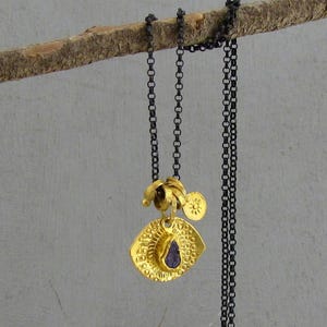May include: A necklace featuring a dark oxidized silver chain and a gold-toned pendant. The pendant consists of a textured, eye-shaped disc with a small purple stone at the center, accompanied by a tiny circular charm embossed with a sun design.