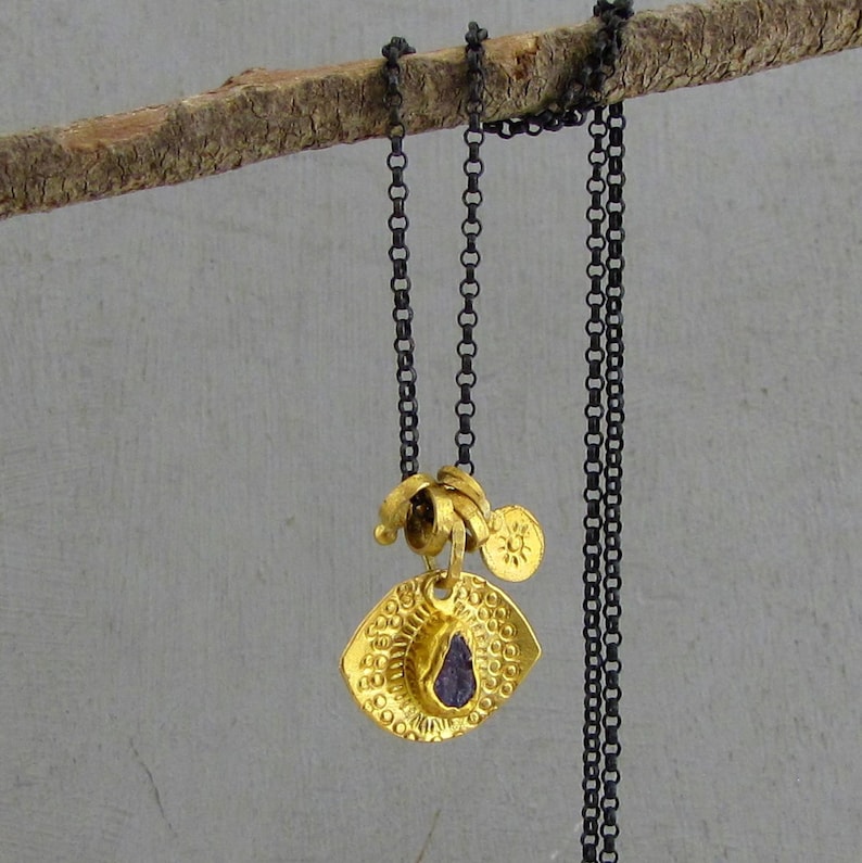 May include: A necklace featuring a dark oxidized silver chain and a gold-toned pendant. The pendant consists of a textured, eye-shaped disc with a small purple stone at the center, accompanied by a tiny circular charm embossed with a sun design.
