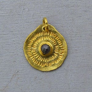 May include: A gold-colored pendant with a central dark gray stone. The pendant has a circular design with concentric lines radiating outwards. It features a small loop at the top for a chain.