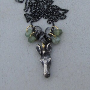 May include: A silver-toned necklace featuring a unicorn pendant with a gold horn. The necklace has a dark chain and is adorned with green gemstone clusters and small gold accents. The unicorn pendant is detailed and textured.