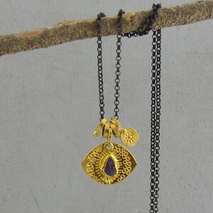 May include: A necklace featuring a dark oxidized silver chain and a gold-toned pendant. The pendant has a textured, eye-shaped base with a small, rough-cut purple stone in the center and a tiny, round gold charm embossed with a sun-like symbol.