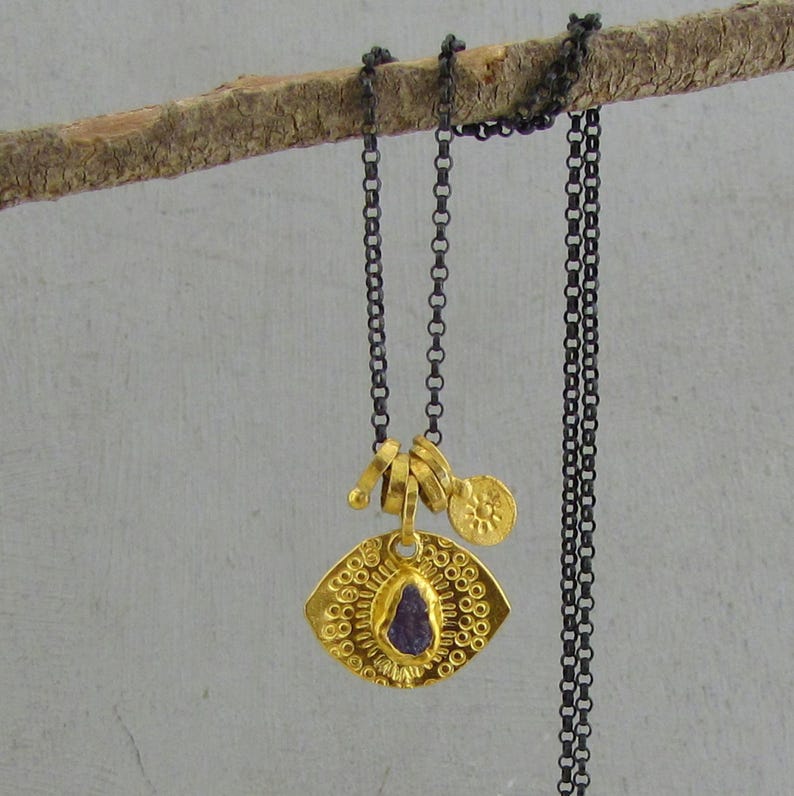 May include: A necklace featuring a dark oxidized silver chain and a gold-toned pendant. The pendant has a textured, eye-shaped base with a small, rough-cut purple stone in the center and a tiny, round gold charm embossed with a sun-like symbol.