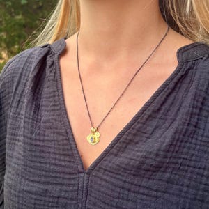 May include: A dark gray chain necklace featuring a small gold-toned pendant with a textured circular base and a central teardrop-shaped blue stone. The pendant includes a tiny gold circular charm attached to the top, resting against a dark gray shirt.