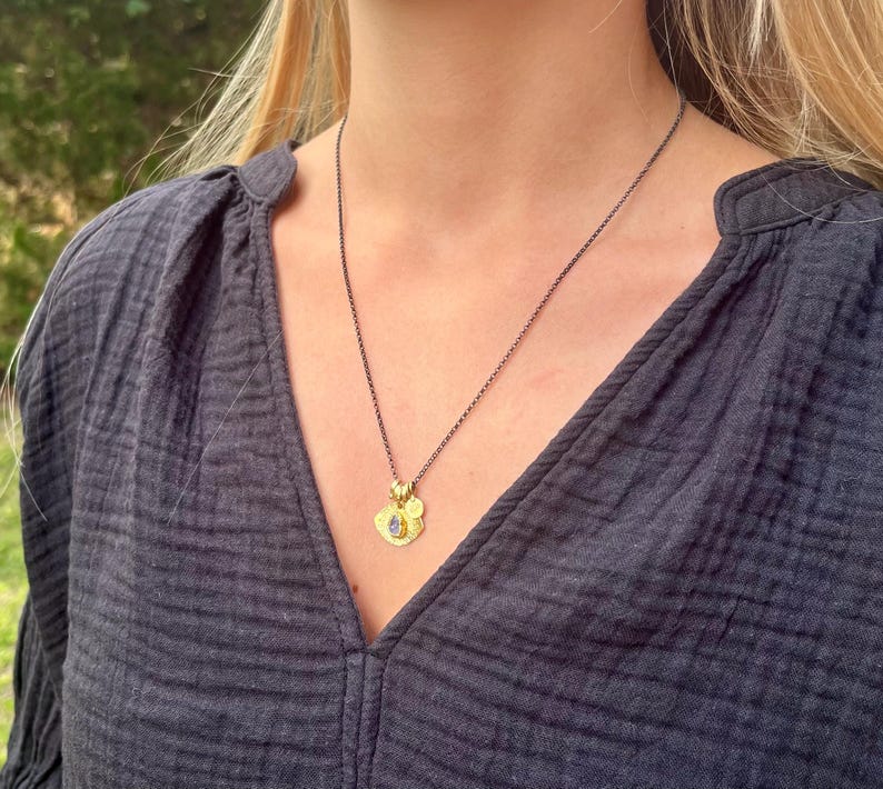 May include: A dark gray chain necklace featuring a small gold-toned pendant with a textured circular base and a central teardrop-shaped blue stone. The pendant includes a tiny gold circular charm attached to the top, resting against a dark gray shirt.