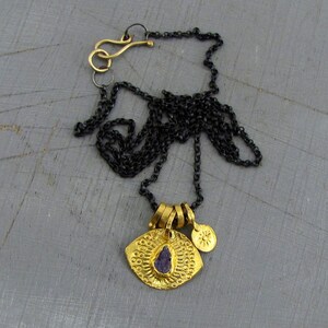 May include: A necklace featuring a dark oxidized silver chain with a gold-toned hook clasp. The pendant consists of a textured gold-toned circular disc with a central purple stone and a small gold-toned charm embossed with a sun-like design.