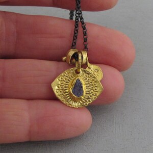 May include: A gold-toned pendant necklace featuring a textured, shield-shaped charm with a central, rough-cut purple stone. The pendant is suspended from a dark, oxidized silver-colored chain and includes a small, circular gold-toned accent tag.
