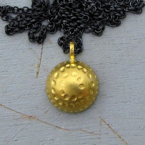 May include: A gold-colored pendant necklace with a textured, round pendant. The pendant is suspended from a dark, oxidized chain. The pendant has a dimpled surface, giving it a unique, handcrafted appearance. The necklace is suitable for everyday wear.