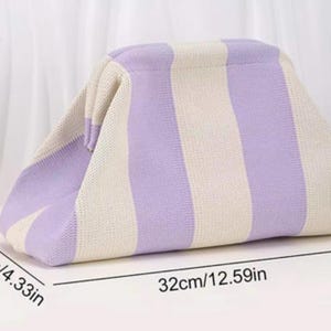May include: A knit clutch bag featuring vertical stripes in light purple and cream. The bag measures 12.59 inches in length, 4.33 inches in width, and 7.08 inches in height. It has a structured frame opening and a soft, textured fabric construction.