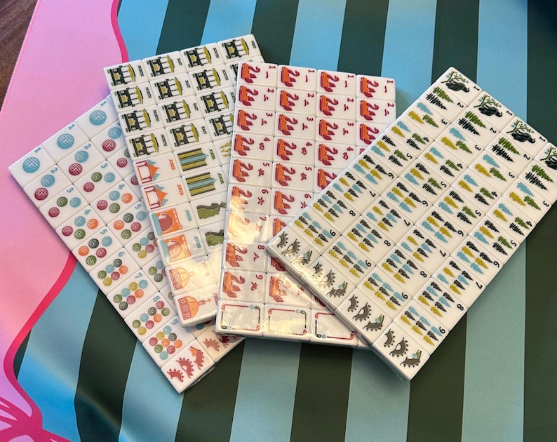 Pu&ograve; includere: Quattro set di tessere Mahjong, ognuno con un design unico. Le tessere sono bianche con simboli e caratteri colorati. I set sono disposti su una superficie a motivi con tonalit&agrave; rosa, verdi e blu.
