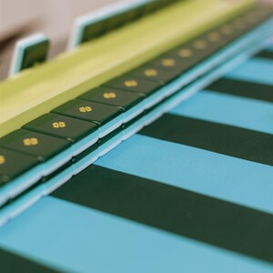 Pu&ograve; includere: Primo piano di un tabellone verde e bianco con tessere verde scuro con simboli dorati. Il tabellone poggia su una superficie a strisce blu e verdi, creando un contrasto visivo accattivante. I pezzi del gioco sono impilati.