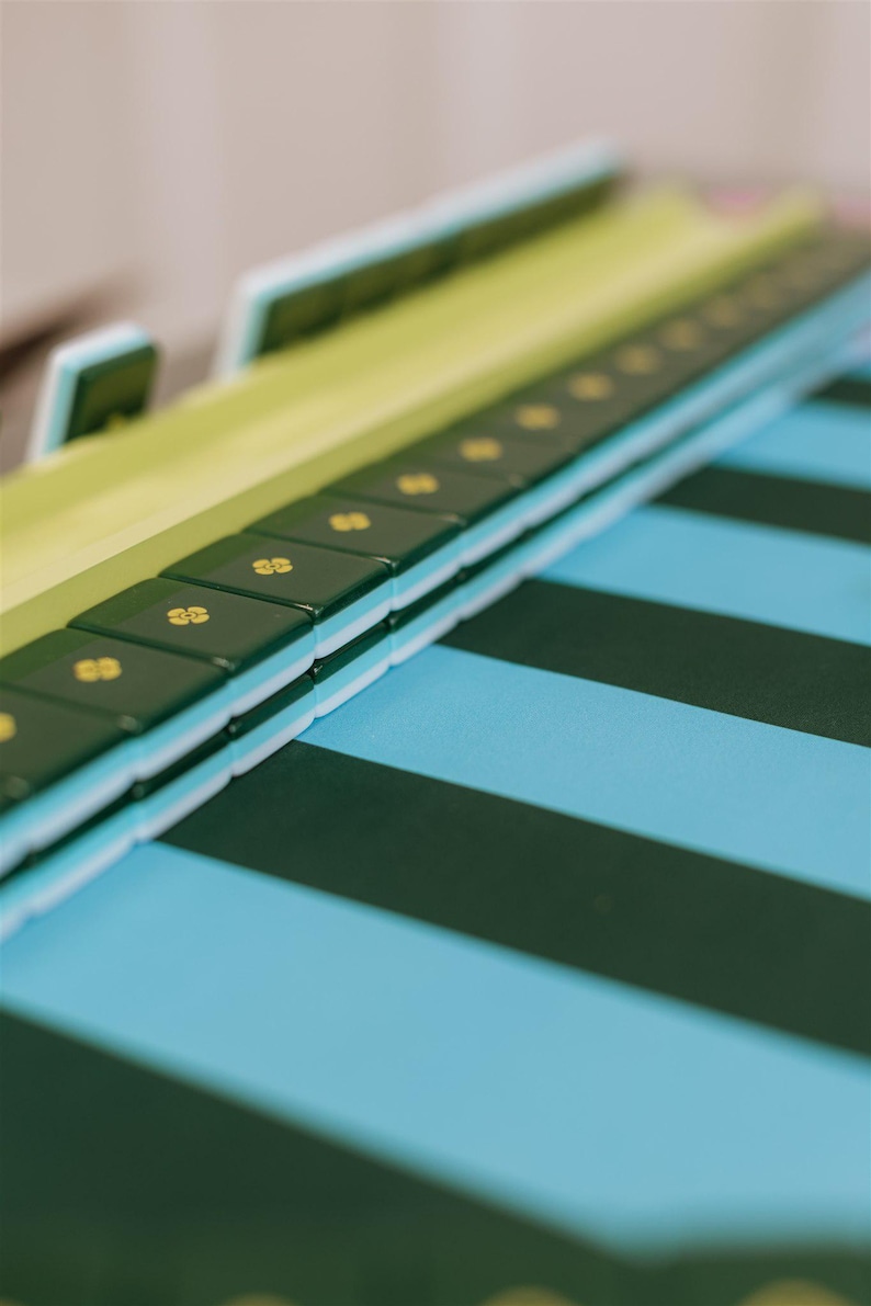 Pu&ograve; includere: Primo piano di un tabellone verde e bianco con tessere verde scuro con simboli dorati. Il tabellone poggia su una superficie a strisce blu e verdi, creando un contrasto visivo accattivante. I pezzi del gioco sono impilati.