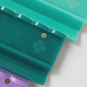 Pode incluir: Tr&ecirc;s suportes retangulares de silicone em tons de verde claro, verde-azulado escuro e roxo. Cada suporte apresenta uma borda superior recortada, um pequeno detalhe met&aacute;lico circular e um design de flor de cinco p&eacute;talas em relevo.