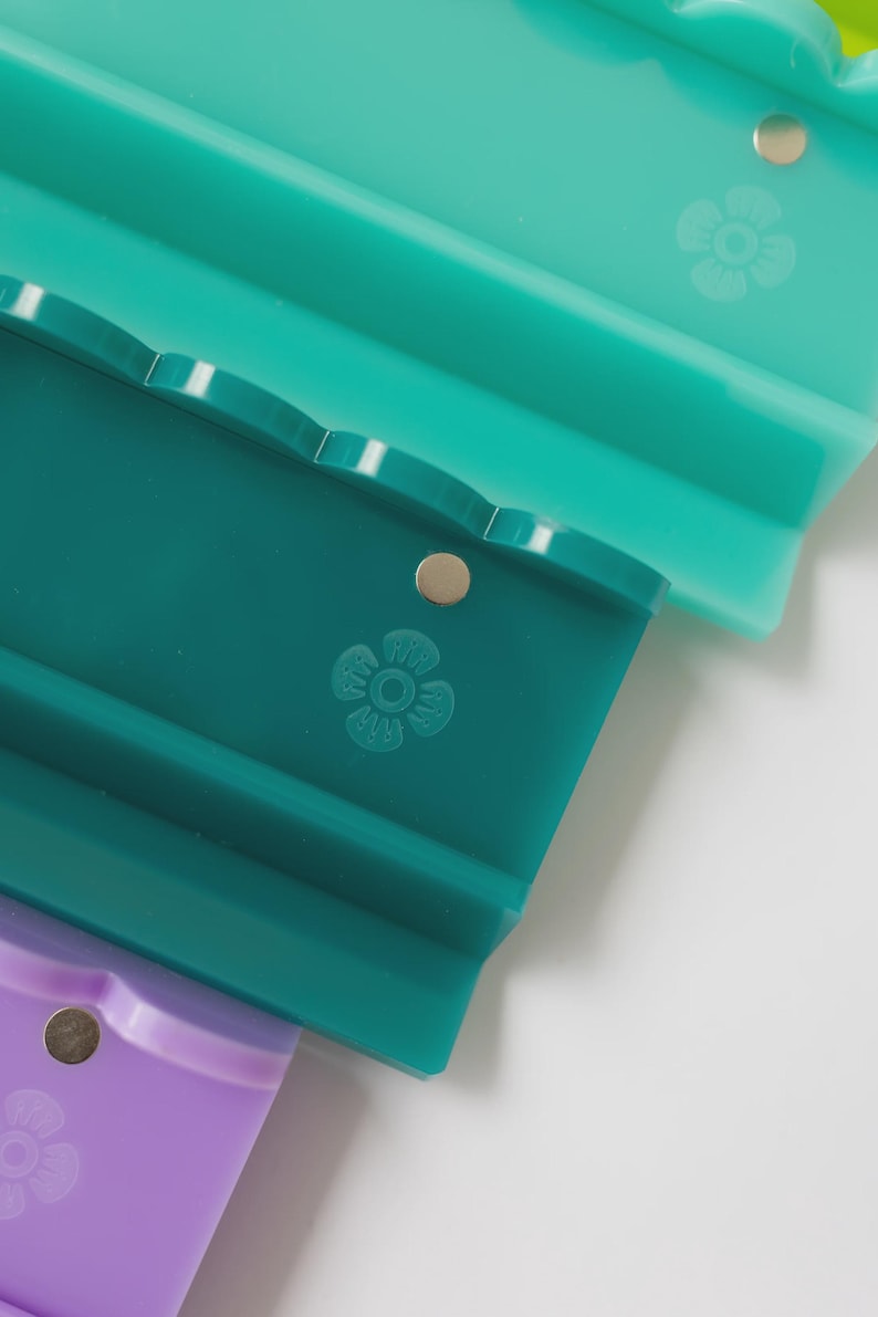 Pode incluir: Tr&ecirc;s suportes retangulares de silicone em tons de verde claro, verde-azulado escuro e roxo. Cada suporte apresenta uma borda superior recortada, um pequeno detalhe met&aacute;lico circular e um design de flor de cinco p&eacute;talas em relevo.