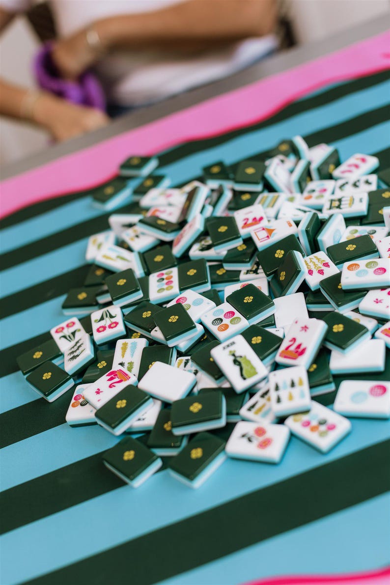 Pu&ograve; includere: Un mucchio di tessere di Mahjong con dorso verde e vari simboli e numeri sulle facce bianche. Le tessere sono sparse su una superficie blu con strisce verde scuro e un bordo rosa. I pezzi del gioco sono a fuoco.