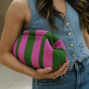 May include: A soft, pouch-style clutch bag featuring a bold, vertical striped pattern in vibrant magenta pink and forest green. The bag has a rounded, gathered shape with a hidden frame closure, constructed from a textured, knit fabric material.