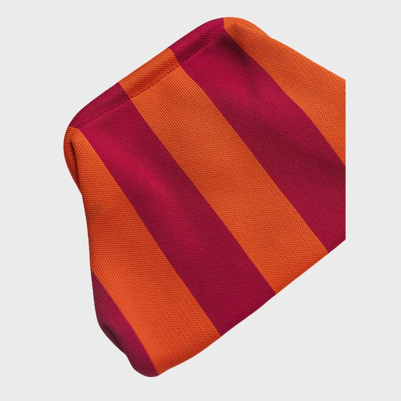 May include: A knitted pouch featuring a repeating pattern of bold, vertical stripes in vibrant orange and deep magenta. The fabric has a consistent, fine-textured weave throughout, with a rounded top edge and a soft, flexible construction.