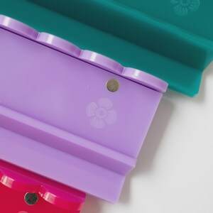 Pode incluir: Tr&ecirc;s suportes retangulares de silicone nas cores azul-petr&oacute;leo, lavanda e magenta. Cada suporte apresenta uma borda superior recortada, um pequeno &iacute;man met&aacute;lico circular embutido perto do centro e um &iacute;cone de flor gravado na base.