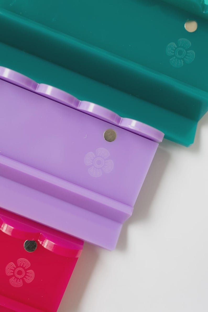 Pode incluir: Tr&ecirc;s suportes retangulares de silicone nas cores azul-petr&oacute;leo, lavanda e magenta. Cada suporte apresenta uma borda superior recortada, um pequeno &iacute;man met&aacute;lico circular embutido perto do centro e um &iacute;cone de flor gravado na base.