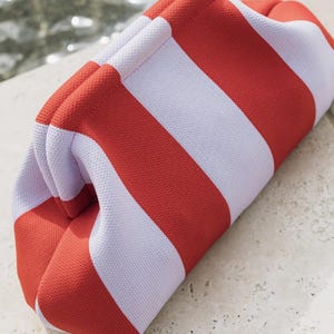 May include: A structured clutch bag featuring a bold, repeating pattern of wide, vertical stripes in vibrant orange and soft white. The bag has a distinct, rounded, frame-style opening with a textured, knit-like fabric construction throughout.