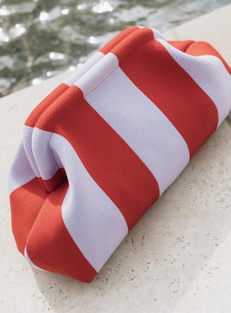 May include: A structured clutch bag featuring a bold, repeating pattern of wide, vertical stripes in vibrant orange and soft white. The bag has a distinct, rounded, frame-style opening with a textured, knit-like fabric construction throughout.