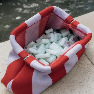 May include: A fabric pouch featuring a bold red and white striped pattern, designed with a wide, open top. The interior is filled with a collection of pale green, rectangular mahjong tiles, each marked with various colorful symbols and characters.