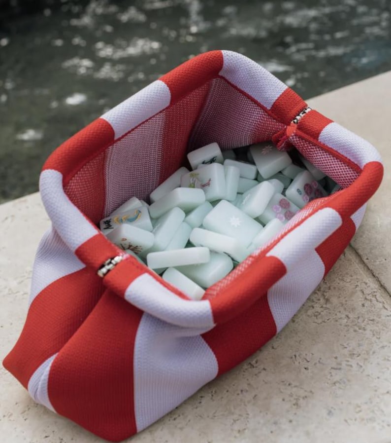 May include: A fabric pouch featuring a bold red and white striped pattern, designed with a wide, open top. The interior is filled with a collection of pale green, rectangular mahjong tiles, each marked with various colorful symbols and characters.