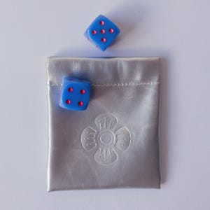 May include: Two blue six-sided dice with pink pips, one showing five and the other showing four, positioned above and beside a silver metallic drawstring pouch featuring a debossed floral emblem centered on the front surface of the bag.