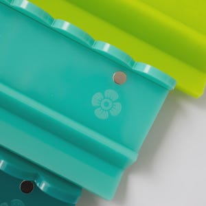 Pode incluir: Um conjunto de tr&ecirc;s tabuleiros retangulares de silicone nas cores verde-lima, azul-petr&oacute;leo e verde-escuro. Cada tabuleiro apresenta um rebordo superior recortado, um pequeno detalhe met&aacute;lico circular e um &iacute;cone de flor gravado na frente.