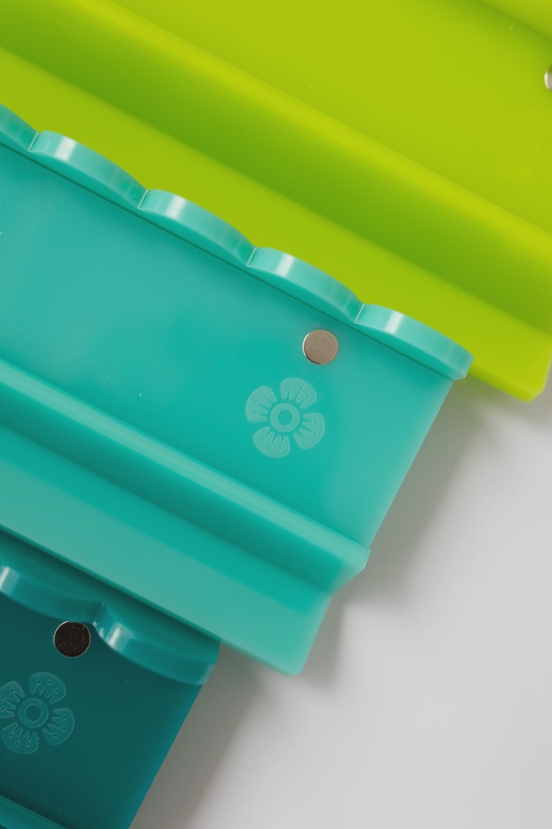 Pode incluir: Um conjunto de tr&ecirc;s tabuleiros retangulares de silicone nas cores verde-lima, azul-petr&oacute;leo e verde-escuro. Cada tabuleiro apresenta um rebordo superior recortado, um pequeno detalhe met&aacute;lico circular e um &iacute;cone de flor gravado na frente.
