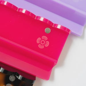 Pode incluir: Tr&ecirc;s ganchos de cabelo retangulares com bordas recortadas, apresentando um pequeno detalhe met&aacute;lico circular e um &iacute;cone de flor em relevo na superf&iacute;cie. Os ganchos s&atilde;o de cor roxa transl&uacute;cida, rosa vibrante e padr&atilde;o de tartaruga castanho.
