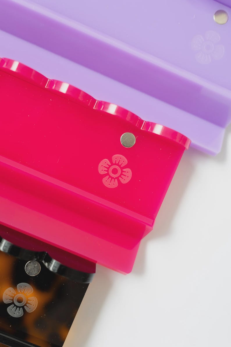 Pode incluir: Tr&ecirc;s ganchos de cabelo retangulares com bordas recortadas, apresentando um pequeno detalhe met&aacute;lico circular e um &iacute;cone de flor em relevo na superf&iacute;cie. Os ganchos s&atilde;o de cor roxa transl&uacute;cida, rosa vibrante e padr&atilde;o de tartaruga castanho.