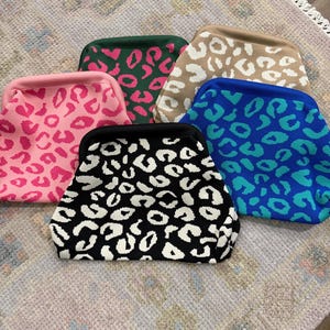 May include: A collection of six knitted clutch bags with a leopard print design. The bags are in various colors, including pink, green, tan, blue, and black, each with a contrasting leopard pattern. The bags are arranged on a patterned rug.