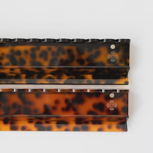 May include: Two rectangular hair accessories with a brown and black tortoiseshell pattern. Each piece features a scalloped top edge, a smooth horizontal ridge, and a small circular cutout near the right side with a flower icon engraved below.