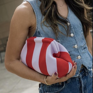 May include: A clutch bag featuring a bold vertical striped pattern in alternating shades of bright red and soft lavender. The bag has a structured, rounded frame with a kiss-lock closure, crafted from a textured fabric material.