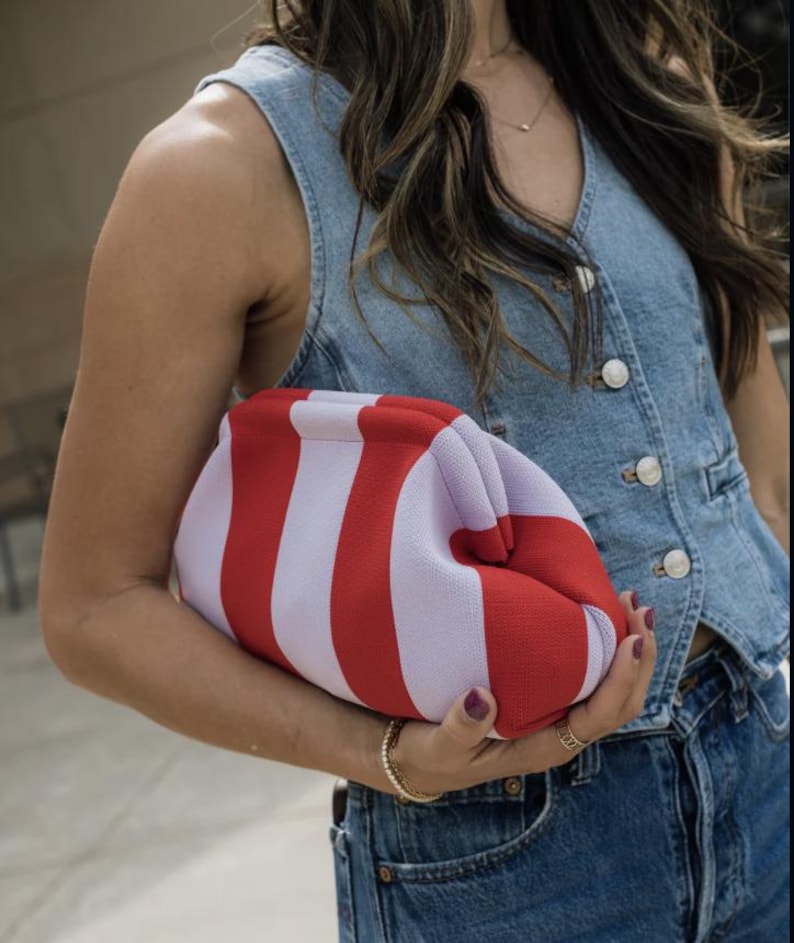 May include: A clutch bag featuring a bold vertical striped pattern in alternating shades of bright red and soft lavender. The bag has a structured, rounded frame with a kiss-lock closure, crafted from a textured fabric material.