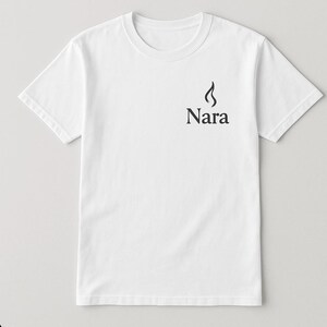 May include: White t-shirt with a crew neck and short sleeves. The shirt features a black graphic of a flame above the word "Nara" printed in a serif font on the upper left chest area. The shirt is made of a soft, lightweight fabric.